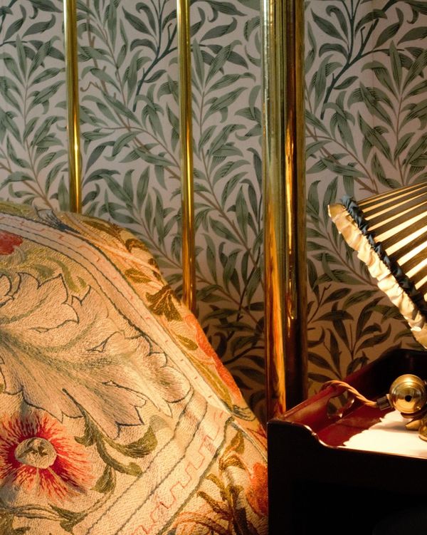 Close detail of green willow-leaf wallpaper behind a brass bed frame and a warmly lit brass lamp
