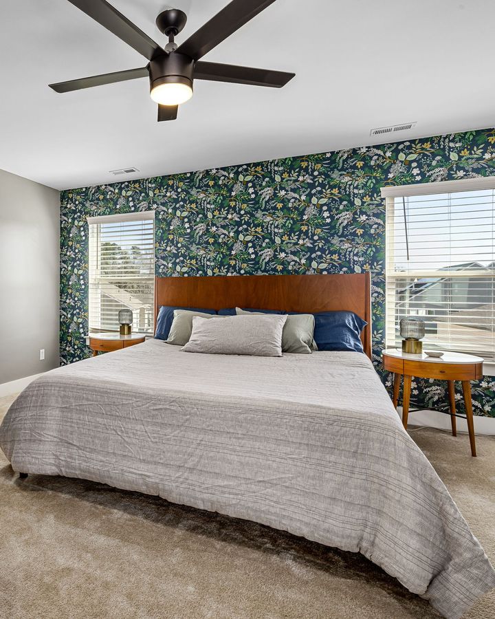 A bedroom with a navy botanical wallpaper accent wall between two windows behind the bed