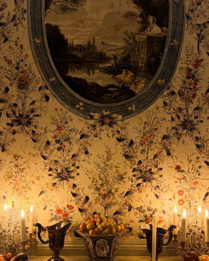 A candlelit dining buffet set against cream wallpaper with fine multicolored florals and an oval framed landscape