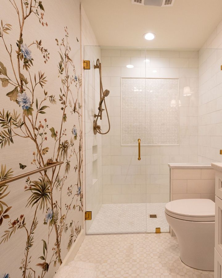 A bright bathroom with chinoiserie branch-and-bird wallpaper, a glass shower and brass fixtures