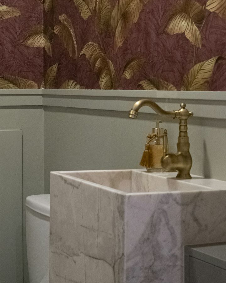 A powder room with plum and gold banana-leaf wallpaper above sage wainscoting, a marble sink and a brass faucet