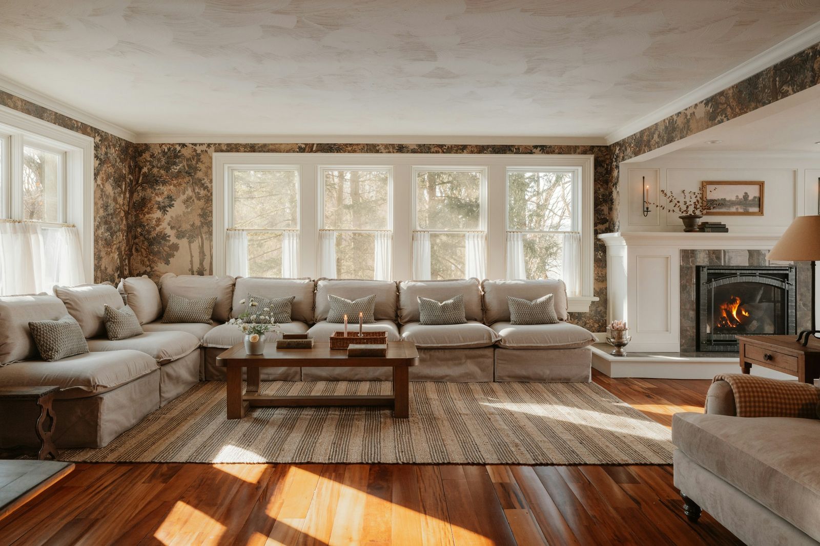 A traditional living room wrapped in scenic forest-mural wallpaper, with white-trimmed windows, a linen sectional and a lit fireplace