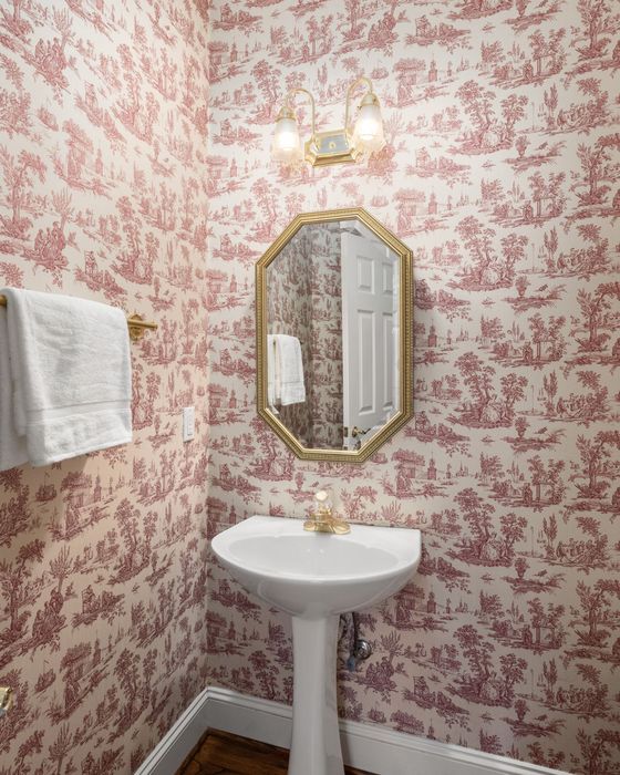 A powder room wrapped in red-on-cream toile wallpaper with a pedestal sink and an octagonal gilt mirror