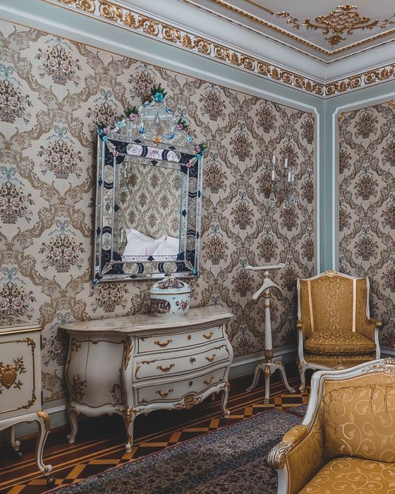 An ornate room papered in a beige and rose floral damask, framed by gilded moldings, with a Venetian mirror and painted commode