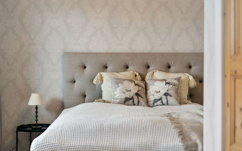 A calm bedroom with soft beige tone-on-tone damask wallpaper behind a tufted linen headboard